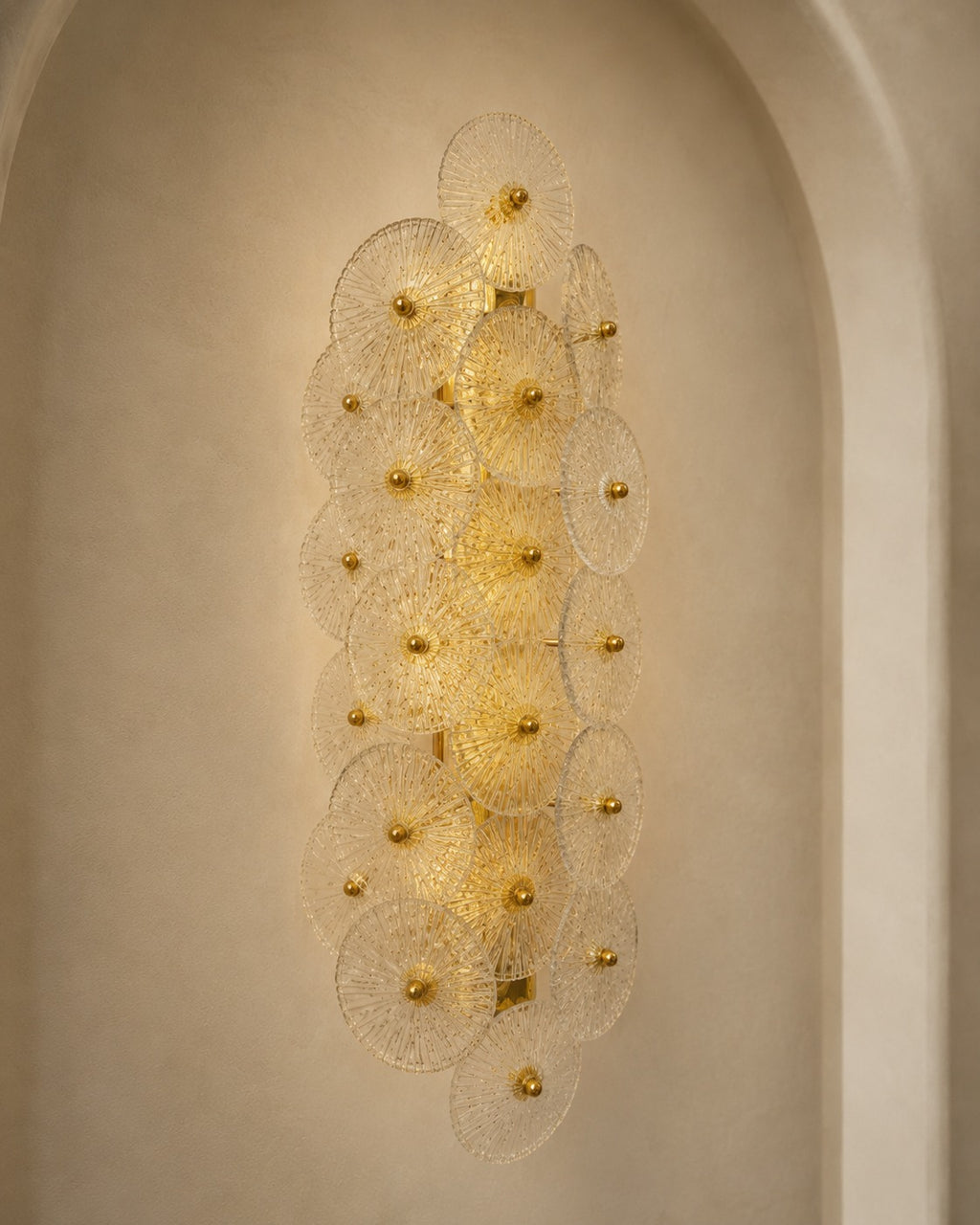 A modern wall sconce light fixture with multiple overlapping circular glass shades and gold-toned central bulbs, mounted on a beige plaster wall in an arched niche.