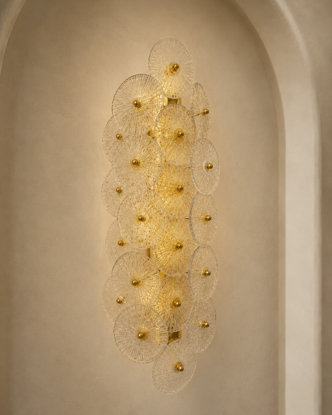 A modern wall sconce light fixture with multiple overlapping circular glass shades and gold-toned central bulbs, mounted on a beige plaster wall in an arched niche.