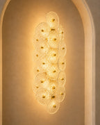 A modern wall-mounted light fixture composed of overlapping circular glass discs with warm gold accents, illuminated against a beige arched wall.