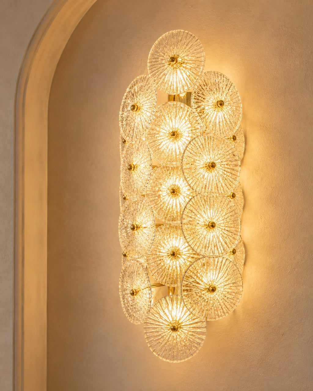 A modern wall sconce featuring a cluster of overlapping textured glass disc elements that glow warmly, mounted on a beige plaster wall with an arched architectural detail.
