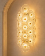 A modern wall sconce featuring a cluster of overlapping textured glass disc elements that glow warmly, mounted on a beige plaster wall with an arched architectural detail.