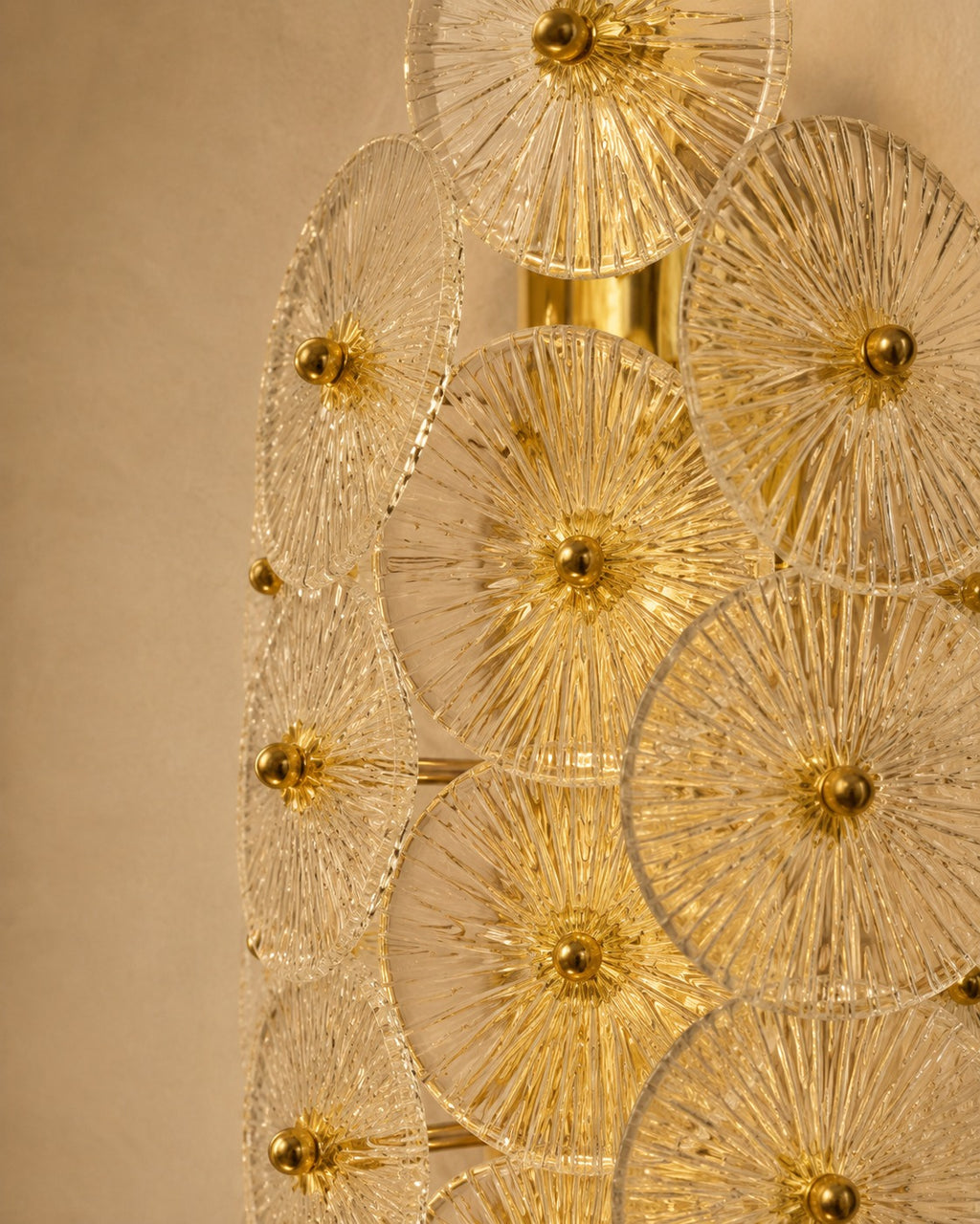 A close-up of a modern wall sconce featuring multiple overlapping clear glass disc elements with radiating gold-toned metal rods and central gold finials, mounted against a warm beige wall.