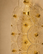 A close-up of a modern wall sconce featuring multiple overlapping clear glass disc elements with radiating gold-toned metal rods and central gold finials, mounted against a warm beige wall.