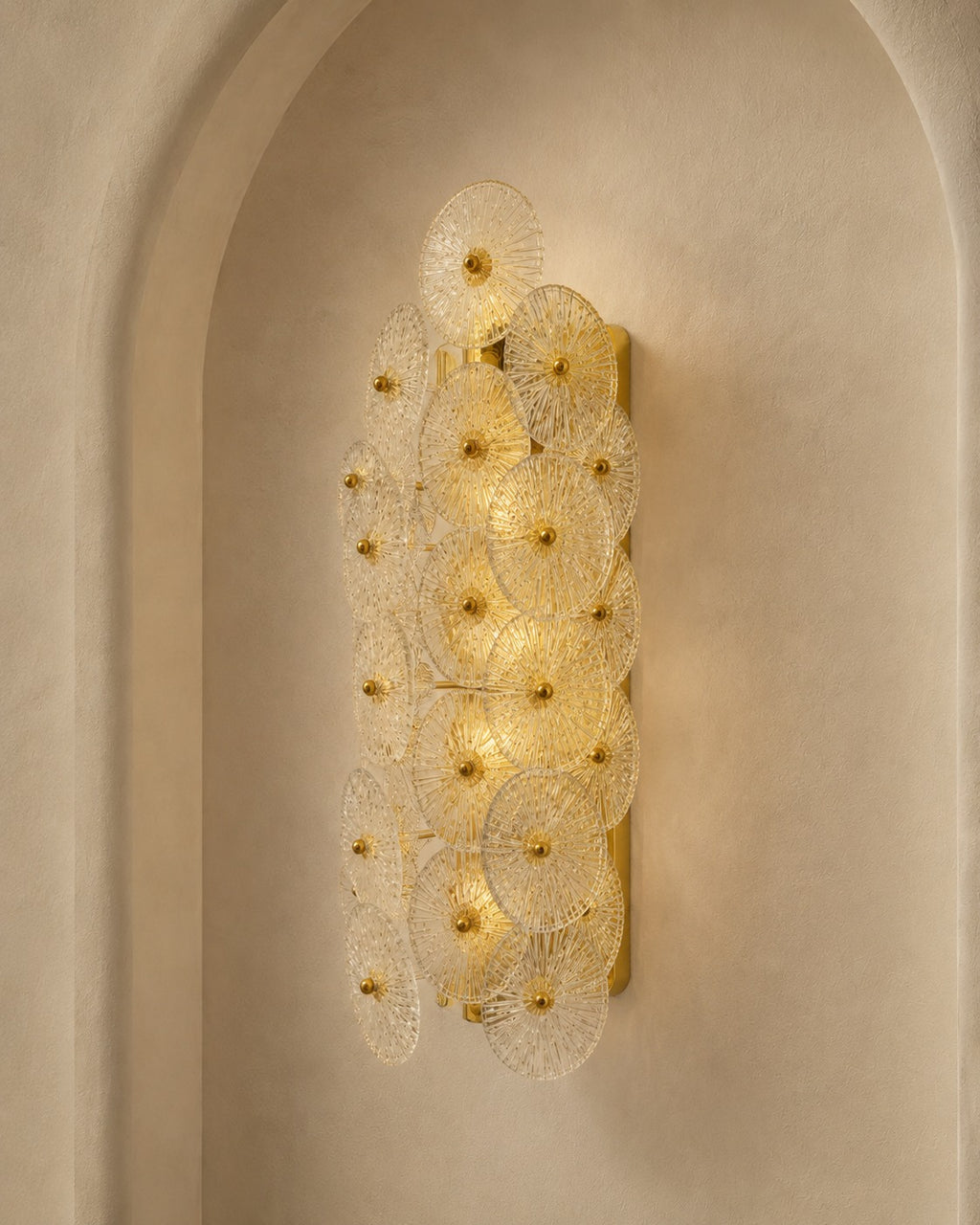 A modern wall sconce light fixture with multiple overlapping circular glass shades and gold-toned metal accents, mounted on a beige plaster wall in an arched niche.