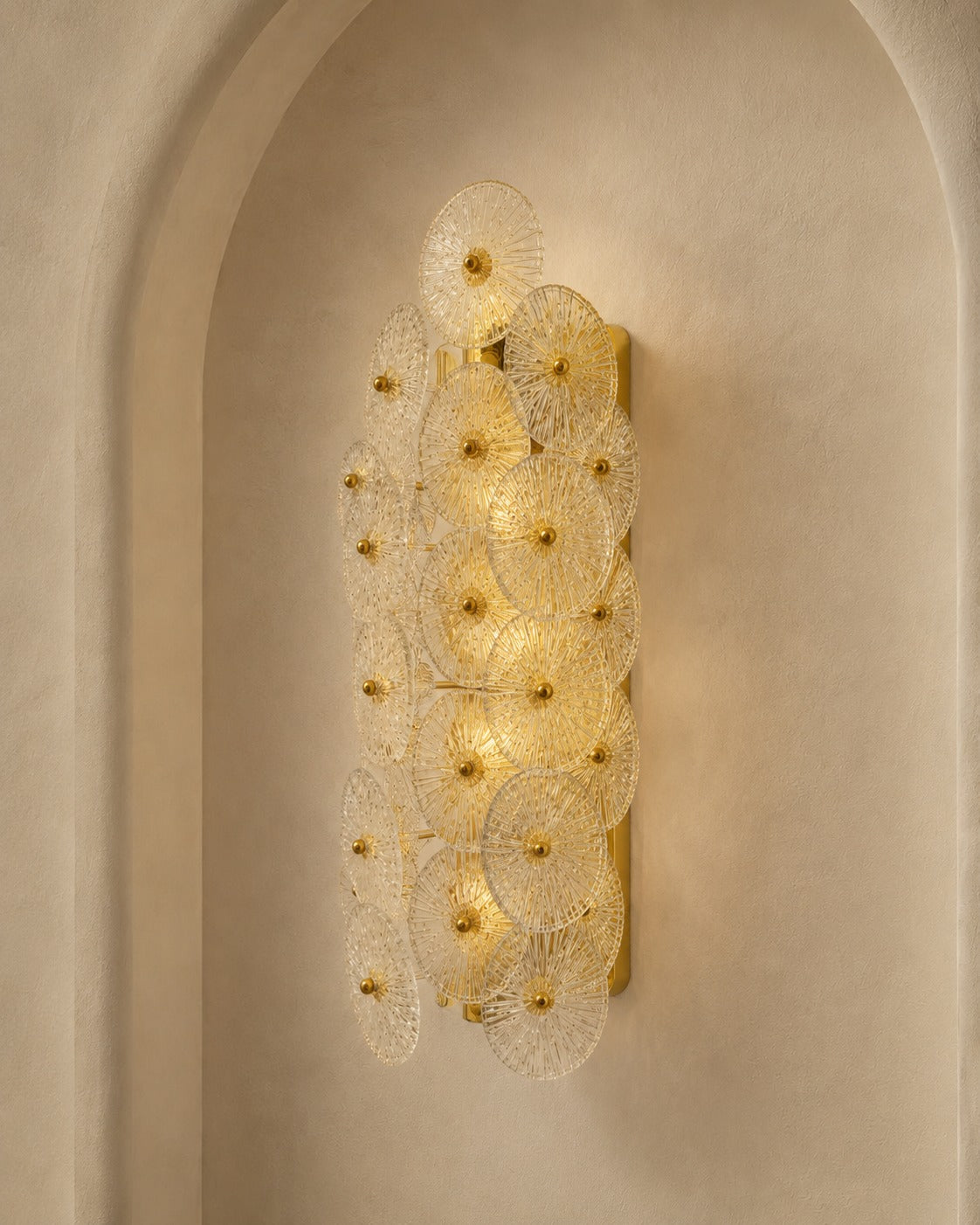 A modern wall sconce light fixture with multiple overlapping circular glass shades and gold-toned metal accents, mounted on a beige plaster wall in an arched niche.